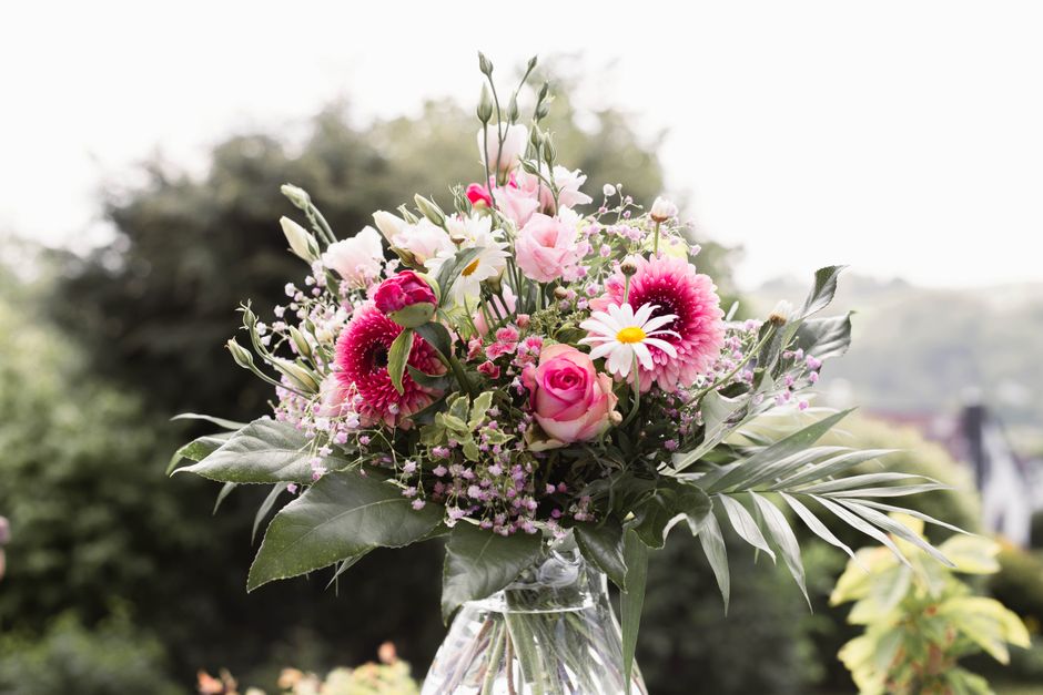 A vibrant floral bouquet arranged in a clear glass vase featuring a variety of fresh flowers including pink roses, white daisies with yellow centers, bright pink gerbera daisies, and delicate pink lis