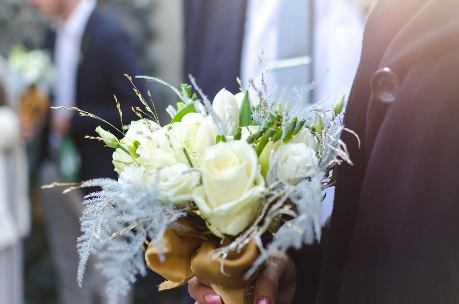 A person holding a floral bouquet featuring white roses, lilies, and delicate greenery, wrapped in elegant golden-brown paper. The bouquet is arranged in a compact, rounded style with fresh, vibrant p