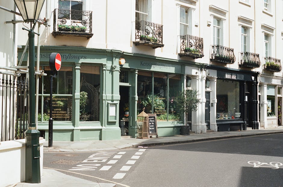 A florist shop with a pastel green storefront, featuring large display windows filled with fresh flowers and plants. The shop has black wrought iron balcony railings on the upper floors of its white-p