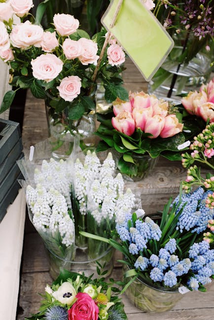 A vibrant display of fresh flowers arranged for delivery, featuring a bouquet of soft pink roses with lush green foliage, placed in a clear glass vase. Nearby, there are soft pink tulips with cream an